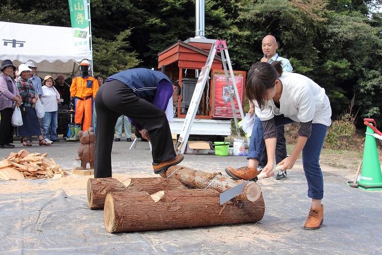 丸太切り大会で女性がハンデをもらい男性と対戦する写真