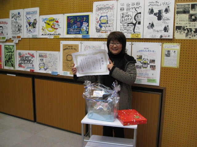自身で手書きされた15年分のポスターをバックに、感謝状と記念品の花を手にする小田川さん