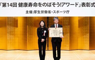 表彰状を手に、前方を向いて立つ西村副市長。その隣には厚生労働省　健康・生活衛生局長　大坪寛子氏。
